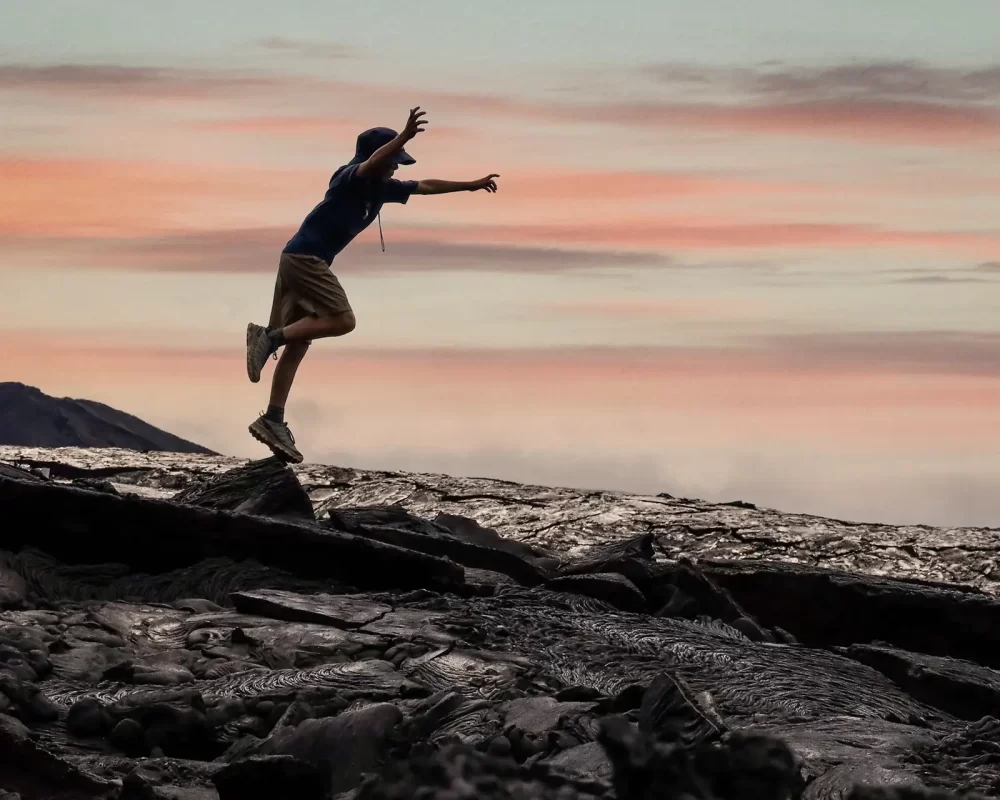 A young boy jumps from a rock while silhouetted against a morning sunrise